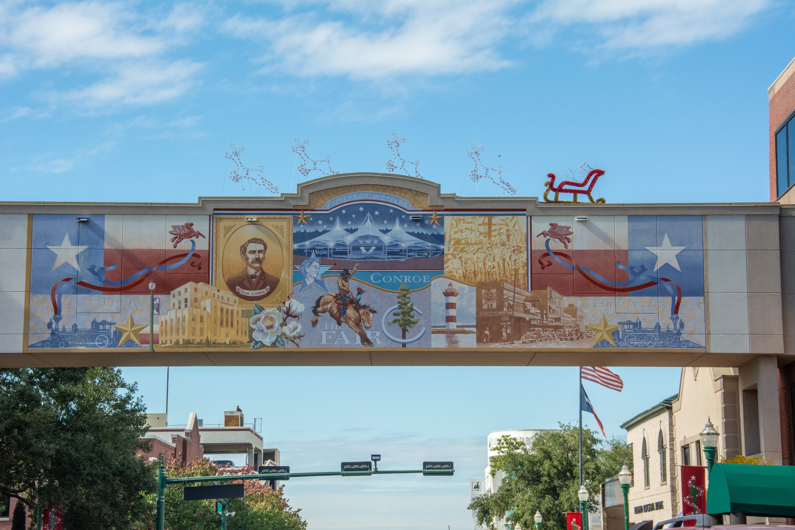 Conroe street view looking up at a historical mural overpass with Texas flags, cowboy art, a train, and Christmas lights.