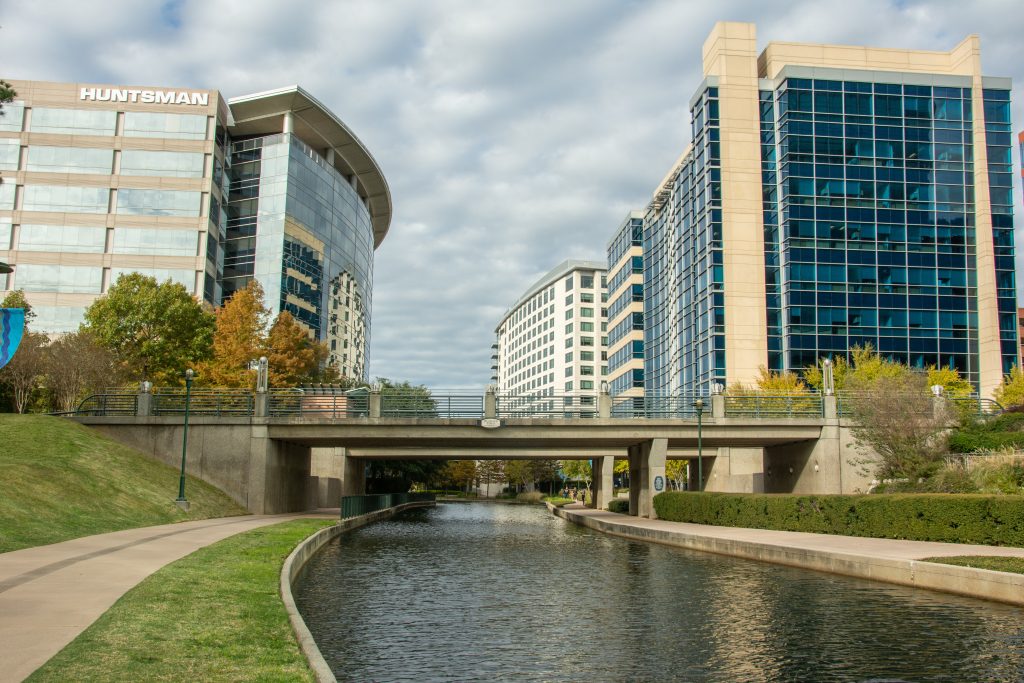 Modern glass corporate high-rise buildings, including the Huntsman building, overlook The Woodlands Waterway, pedestrian bridge, and walking path under a cloudy sky in The Woodlands, Texas.